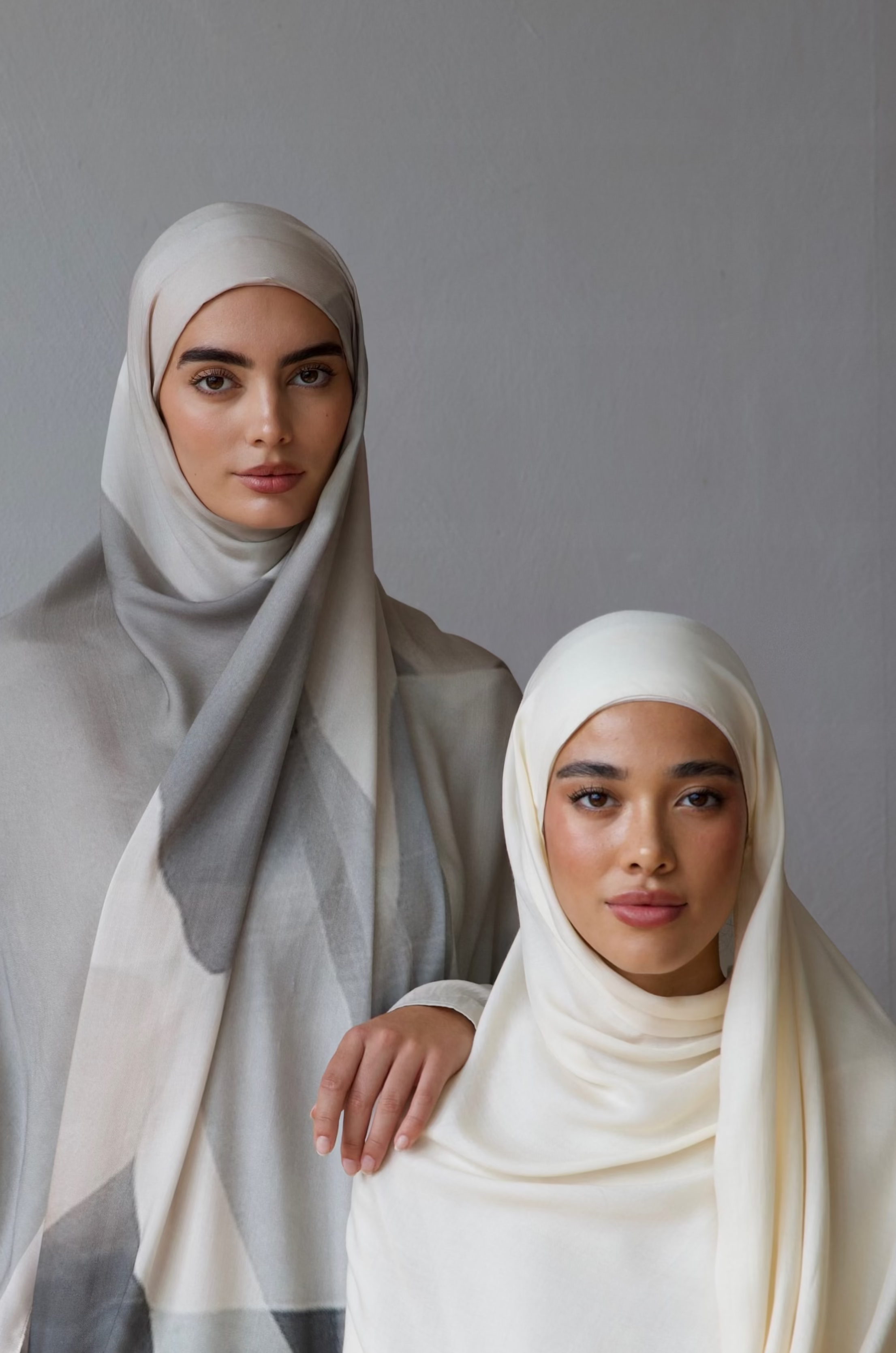 Two women wearing hijabs standing against a plain background