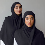 Two women wearing dark navy grey hijabs draped over white outfits, standing against a plain light gray wall.