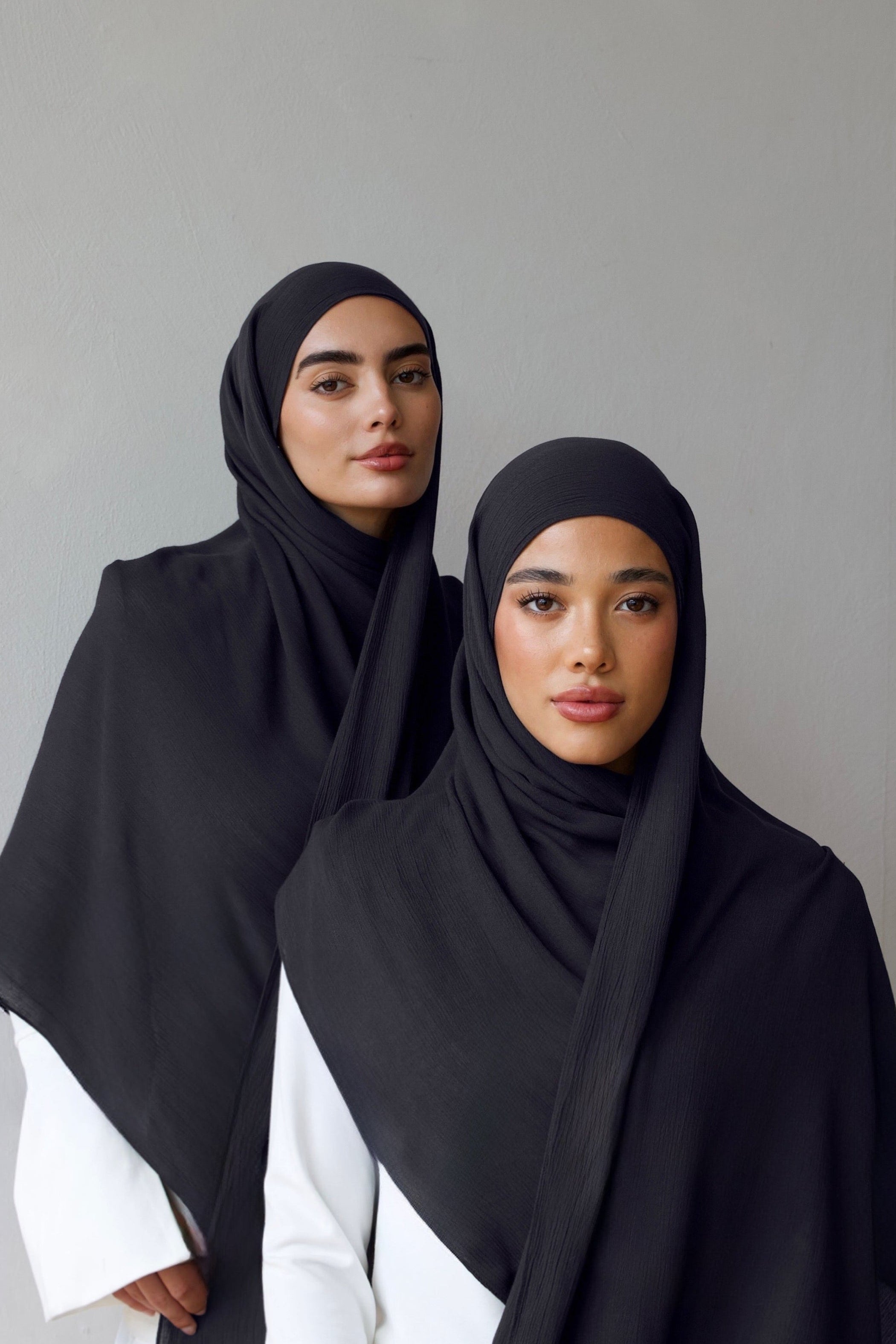 Two women wearing dark navy grey hijabs draped over white outfits, standing against a plain light gray wall.