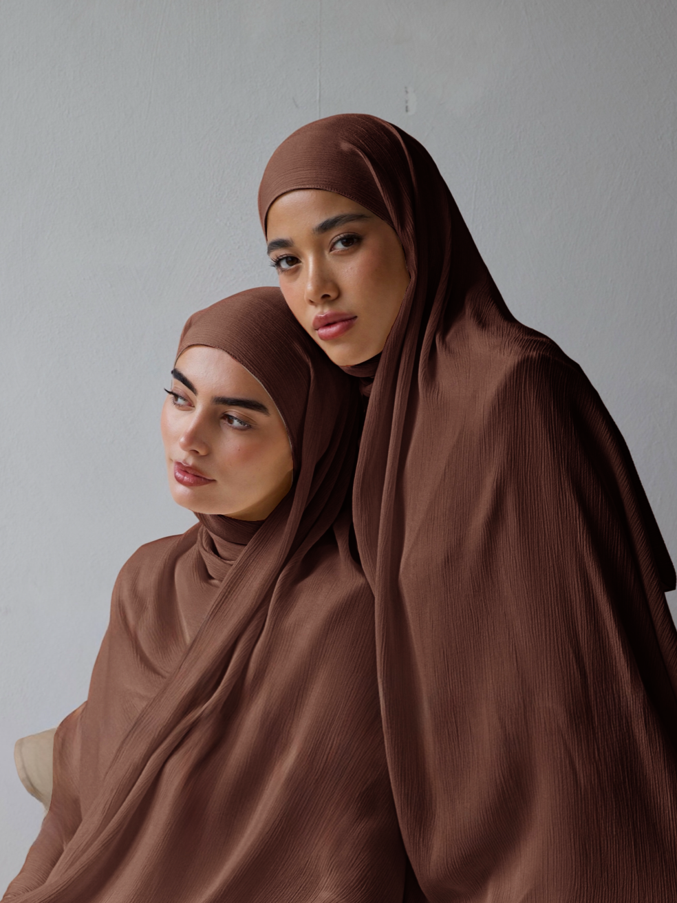 Two women wearing brown hijabs sitting on a gray background