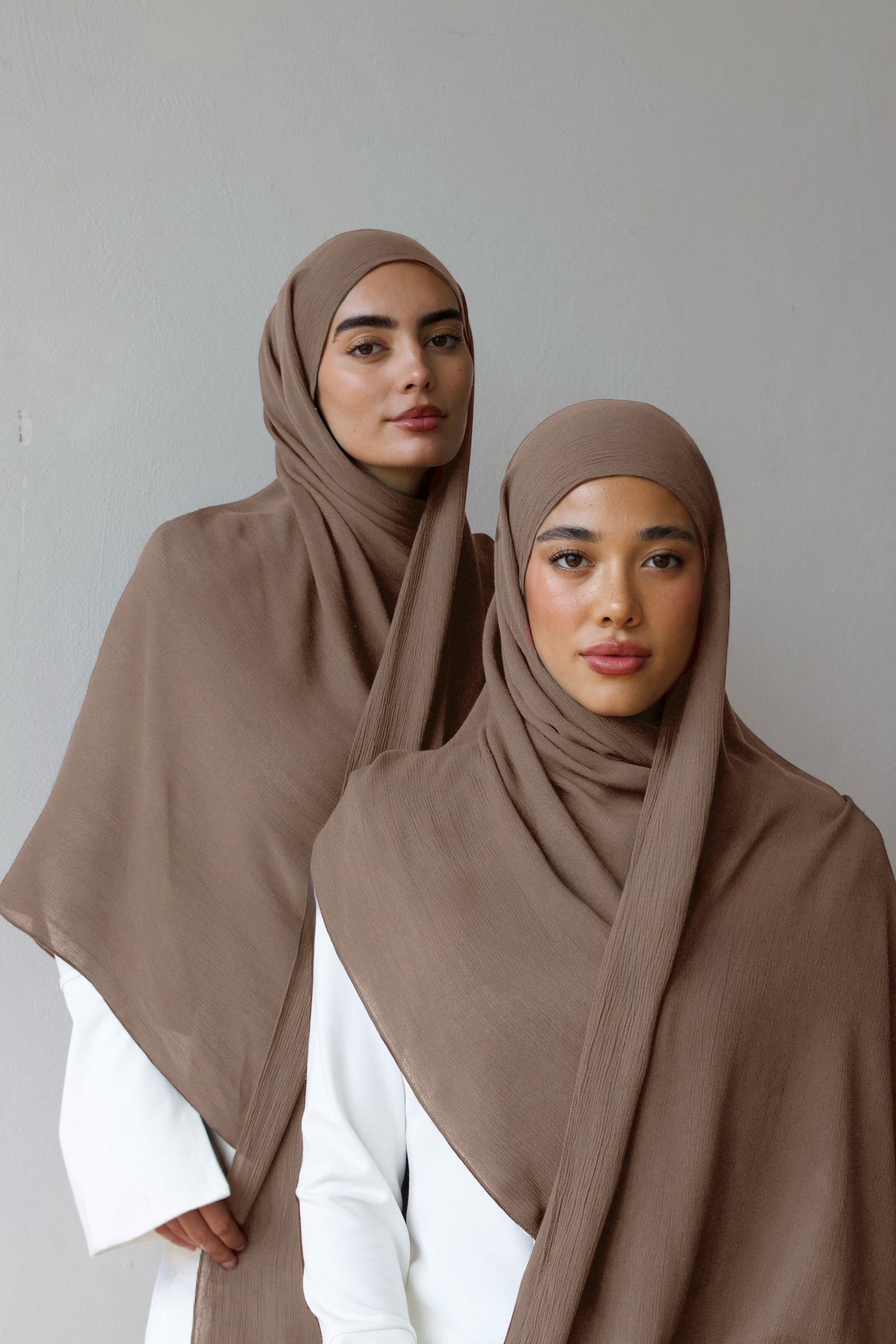 Two women wearing brown hijabs and white outfits against a gray background