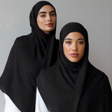 Two women modeling black hijabs and shawls over white outfits, standing against a plain light gray wall.