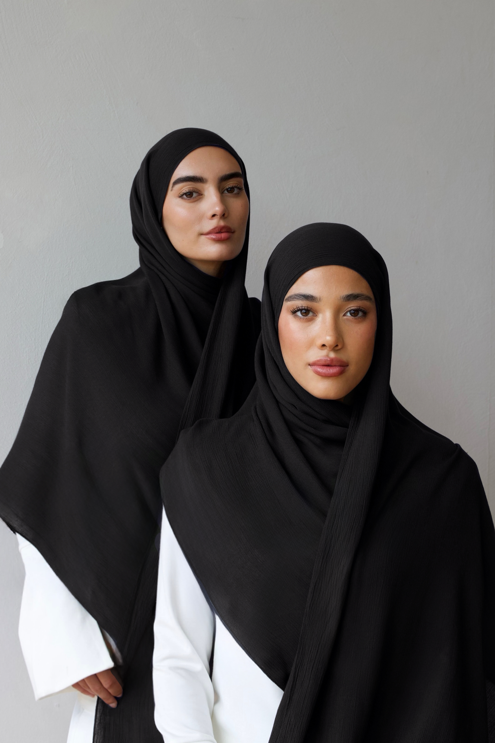 Two women modeling black hijabs and shawls over white outfits, standing against a plain light gray wall.