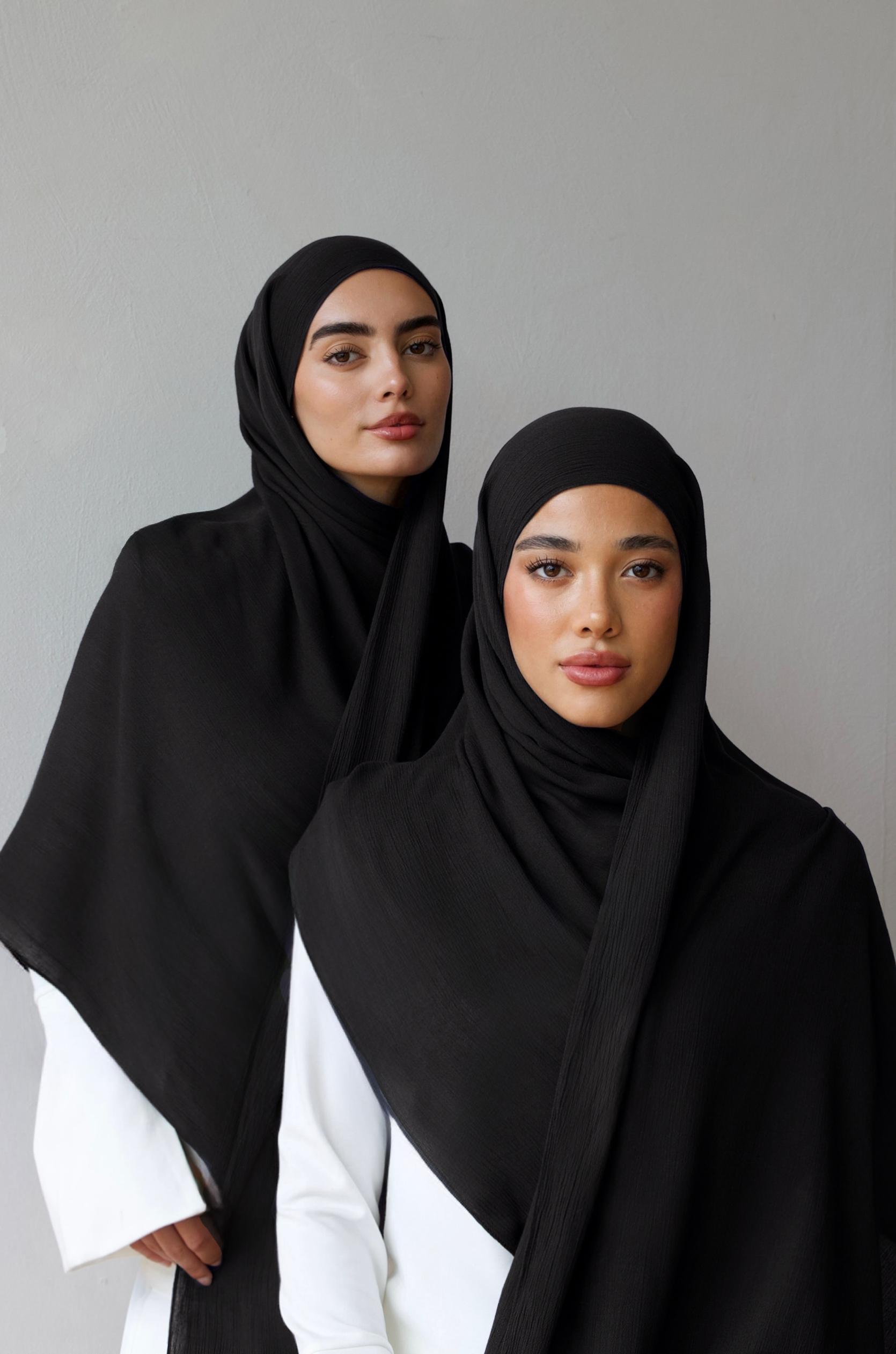 Two women modeling black hijabs and shawls over white outfits, standing against a plain light gray wall.