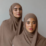 Two women wearing matching taupe-brown hijabs draped over white tops, posed against a plain light gray studio background.