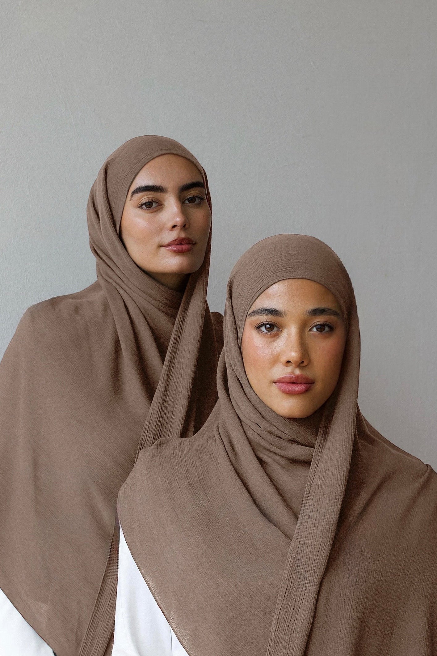 Two women wearing matching taupe-brown hijabs draped over white tops, posed against a plain light gray studio background.