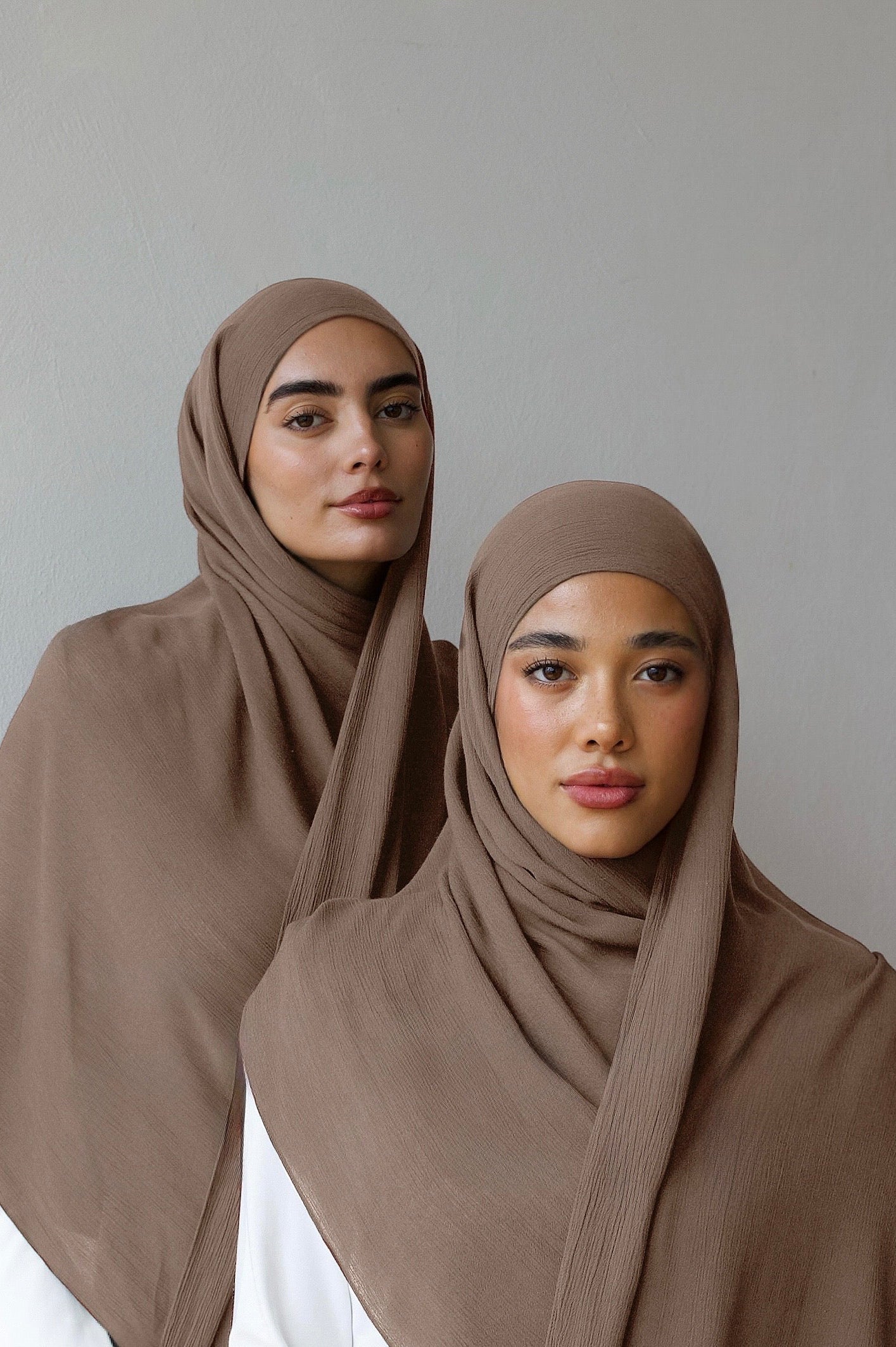 Two women wearing matching taupe-brown hijabs draped over white tops, posed against a plain light gray studio background.