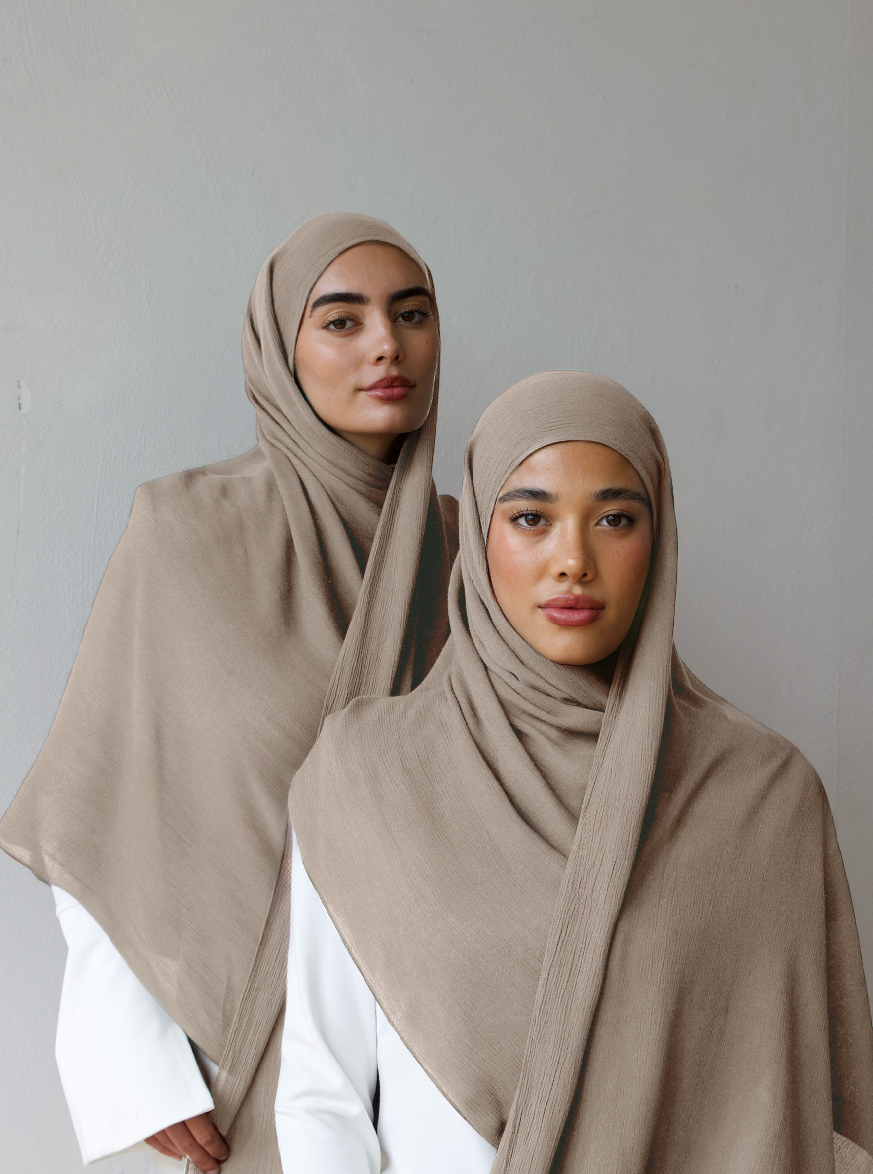 Two women wearing beige hijabs and white clothing against a plain background