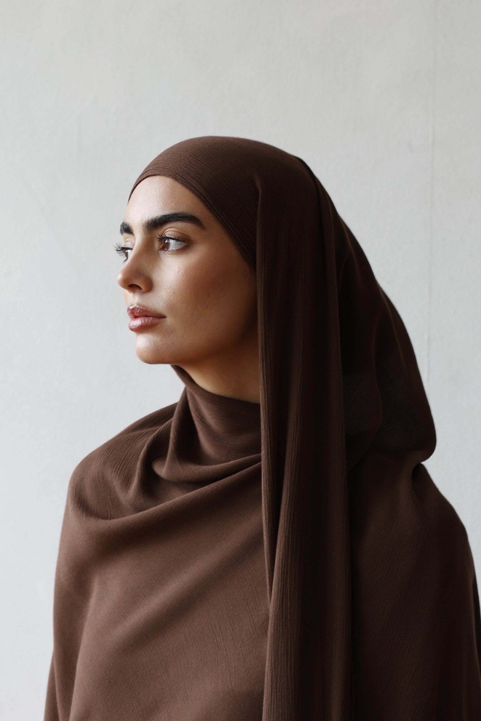Woman wearing a brown hijab and white outfit against a plain background