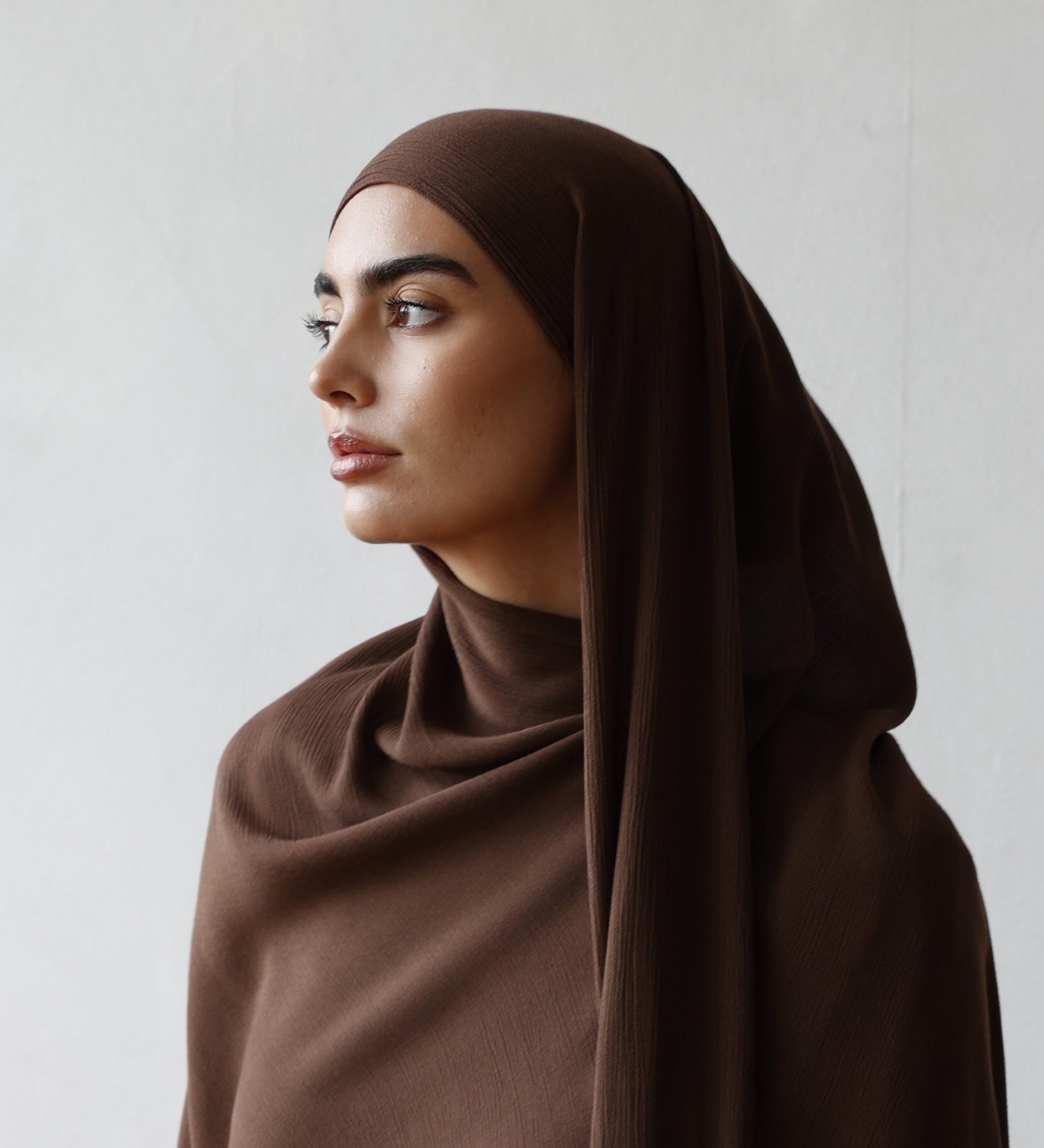 Woman wearing a brown hijab and white outfit against a plain background