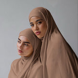 Two women wearing matching taupe brown hijabs and loose draped shawls, posed against a plain light gray studio background.