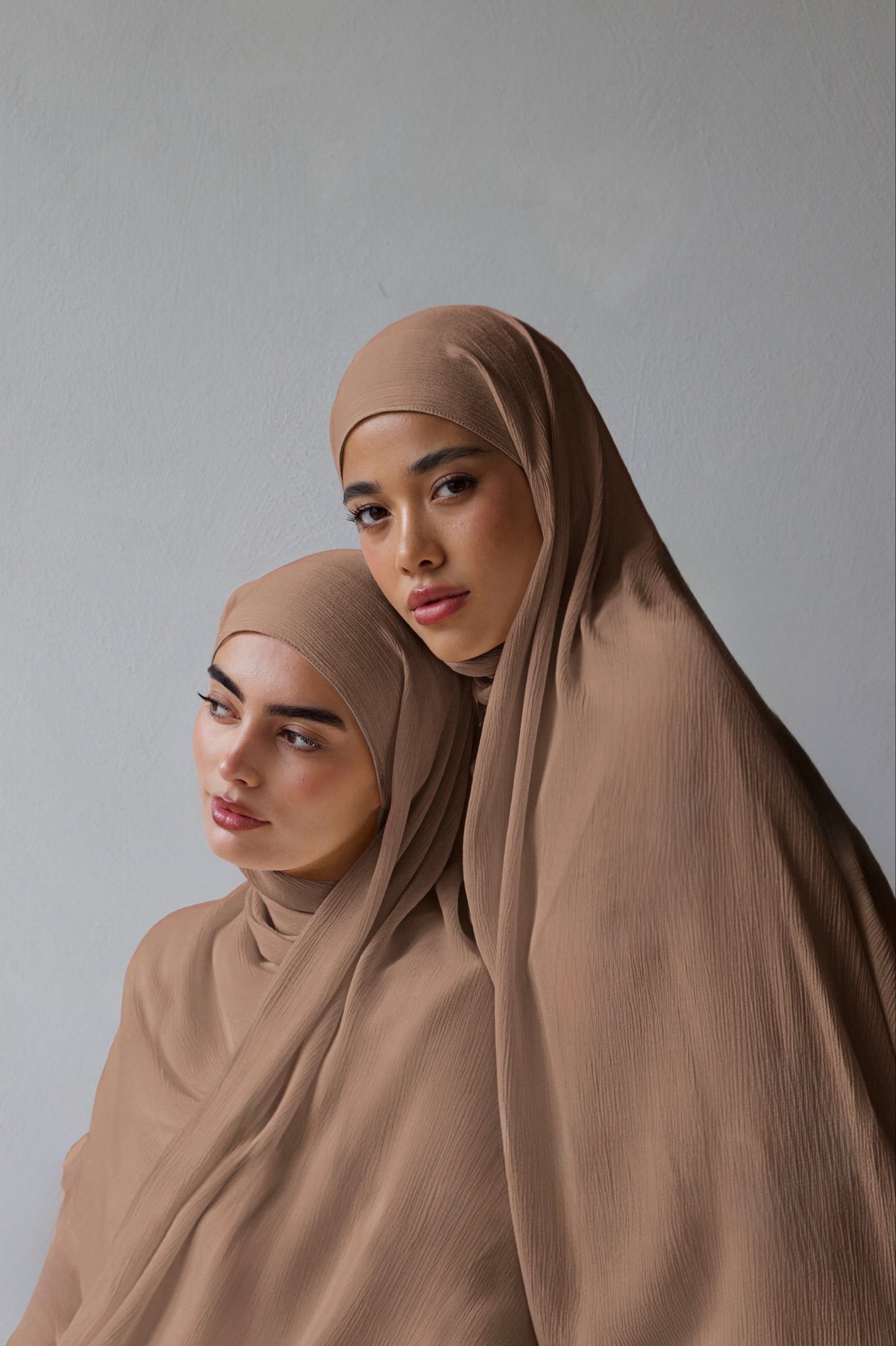 Two women wearing matching taupe brown hijabs and loose draped shawls, posed against a plain light gray studio background.